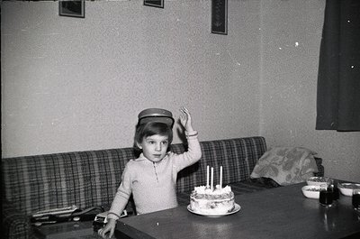 Child in 1960s-70s indoor setting, seated at a wooden table with a small birthday cake (three lit candles) and floral pattern...