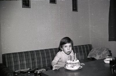 Vintage black-and-white photo of a young child seated at a table, blowing out candles on a round birthday cake. Mid-century i...