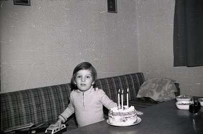 Vintage black-and-white photo of a child seated at a wooden table, blowing out three lit candles on a round cake. Plaid sofa ...