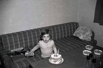 Black-and-white snapshot of a child seated on a plaid sofa, holding a toy vehicle while staring at a small birthday cake with...