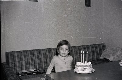 Vintage black-and-white photo of a child seated at a table with a three-candle birthday cake. Plaid sofa and worn wallpaper s...