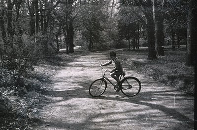 A child rides a classic single-speed bicycle on a gravel path through a wooded area, framed by dense trees. Mid-20th century ...