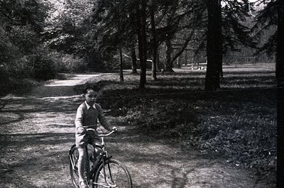 Mid-20th century black-and-white photo of a child riding a simple bicycle on a rural gravel path, surrounded by dense trees a...