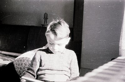 Mid-20th century black-and-white portrait of a young child seated indoors, wearing a patterned sweater and dark trousers. The...