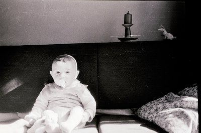 Vintage black-and-white photo of an infant seated on a bed, holding a toy. Simple wooden nightstand with a candle holder and ...
