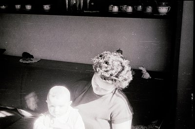 Vintage black-and-white photo of two children playing on a cluttered floor with a shelf of white ceramic mugs in background. ...