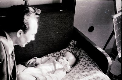 A man in mid-20th-century attire cradles a sleeping infant in a hospital bassinet, likely post-WWII or Cold War era. Floral-p...