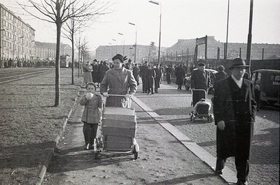 Mid-20th century urban street scene with families pushing strollers, likely post-WWII. Concrete sidewalks, bare trees, and a ...