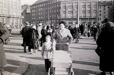 Mid-20th century urban street scene with Soviet-style architecture. Crowded plaza featuring a woman pushing a baby stroller, ...