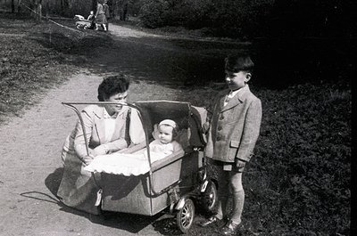 Mid-20th century family stroll in a rural park: woman in a long coat pushes a vintage pram with a baby, while a young boy in ...