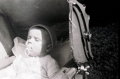 Vintage black-and-white photo of an infant sleeping in a crib with a pacifier, dressed in a simple onesie. Partial view of a ...