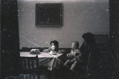 Vintage sepia-toned indoor scene featuring three seated figures at a table with a cake. Adults and child in formal attire, li...