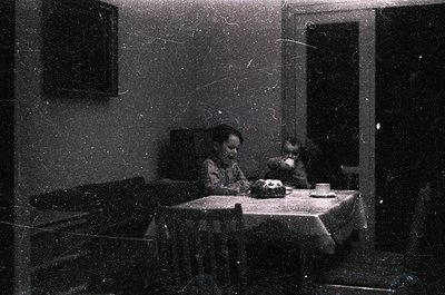 Vintage black-and-white interior shot of two children seated at a small table with a cake, teacup, and cloth napkin. Plain wa...
