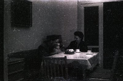 Vintage sepia-toned indoor scene featuring two individuals seated at a small table with a cake and cups. Man in dark cap, wom...