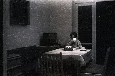 Vintage black-and-white interior shot of a child seated at a round wooden table with a white tablecloth, holding a cup. Sideb...