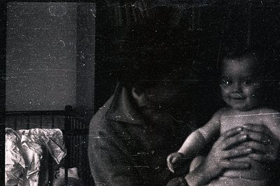 Vintage black-and-white photo of an infant cradled in an adult’s arms, likely mid-20th century. Soft focus and grainy texture...