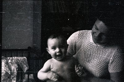 Vintage black-and-white photo of a woman cradling a toddler indoors, likely mid-20th century. The woman wears a patterned blo...