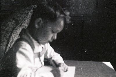 Vintage black-and-white photo of a young child engrossed in writing at a small wooden desk, wearing a white blouse with a lac...