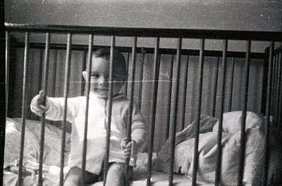 Vintage black-and-white photo of a child in a metal-framed crib, giving a thumbs-up. Simple striped onesie and blanket visibl...