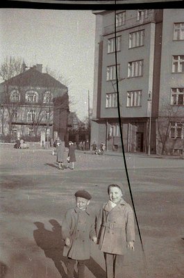 Two children in 1950s-60s Eastern European streetwear pose in front of Soviet-era concrete apartment block. Mixed architectur...