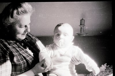 A vintage black-and-white photo of an elderly woman holding a baby indoors, likely mid-20th century. The woman wears a plaid ...