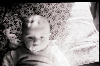 Vintage black-and-white photo of an infant in a floral-patterned blanket, holding a pacifier. Soft focus and grainy texture s...