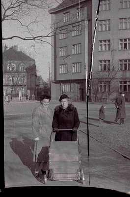 Two women in 1950s-era clothing pose with a baby carriage on a paved urban street flanked by mid-rise brick buildings. One we...