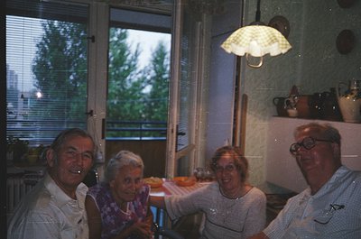Four adults pose indoors near an open window overlooking greenery and distant city lights. Mid-century kitchen features a pen...