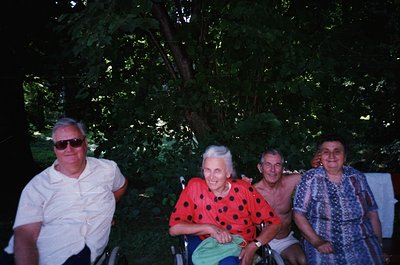 Four adults pose outdoors under dense foliage, likely in a garden or park setting. The man on the left wears sunglasses and a...
