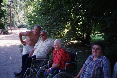 Four seniors pose outdoors in a shaded park setting, likely mid-20th century. Two men stand—one shirtless, one in a light-col...