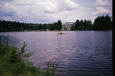 Vintage lakeside scene featuring a multi-story lakeside resort with classic 20th-century architecture. A single kayaker paddl...
