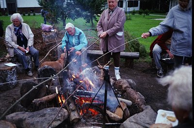 Group of five adults skewering food over an open fire in a grassy outdoor setting, likely mid-20th century. Men in suits and ...