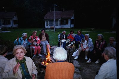 Group of 12+ adults gathered around an outdoor fire pit in a residential backyard, 1970s-1980s. Casual attire includes sweate...