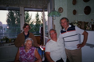Four adults pose indoors in a mid-century kitchen, likely 1970s–1980s. Light-colored walls, checkered curtains, and ceramic p...