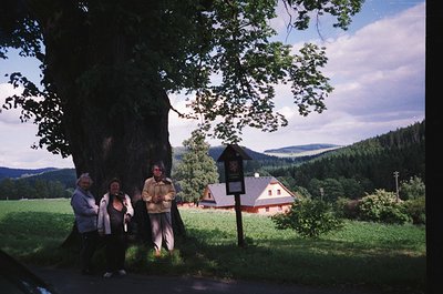 Three individuals pose under a large tree beside a vintage signpost in a rural landscape. The signpost displays a red-and-whi...