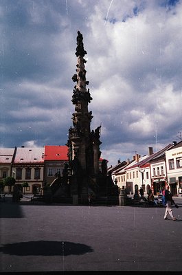 Baroque-style column monument with intricate sculptures, likely a 17th–18th century urban square. Surrounding buildings featu...