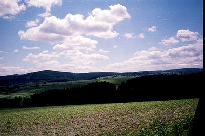 Vintage landscape shot of rolling farmland under bright, fluffy clouds. Green fields stretch toward forested hills under clea...