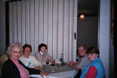 Vintage indoor gathering of five adults seated at a round table in a dimly lit room with vertical blinds. Mid-century retro d...