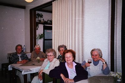 Vintage group portrait of five elderly women in a dimly lit indoor setting, likely a 1980s-1990s European home. Seated at a t...