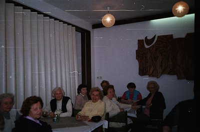 Vintage indoor gathering in a minimalist 1970s-style café or lounge. Eight adults seated around a rectangular table, dressed ...