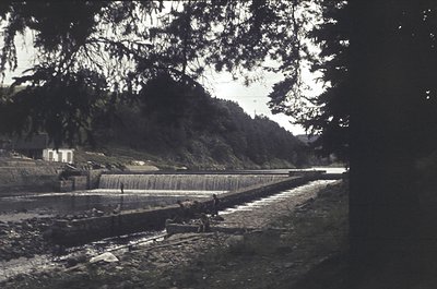 Vintage sepia-toned photo of a small waterfall cascading into a shallow riverbed, framed by dense forest. A lone figure sits ...