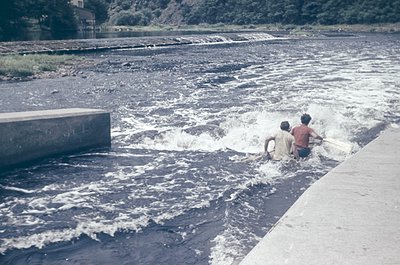 Two young boys wade through turbulent water at a concrete dam spillway, mid-1960s. Vibrant greens contrast with the concrete ...