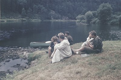 Five individuals relax by a serene lake, seated on grass beside a canoe. Mid-20th century clothing and vintage camera suggest...