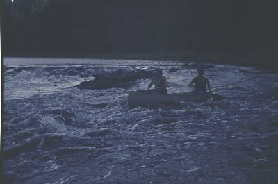 Two individuals paddle a canoe through turbulent waters under low light, likely dawn/dusk. Vibrant blue-green hues dominate t...