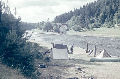 Vintage sepia-toned scene of a riverside campsite with three tents pitched near a small wooden house. Two individuals stand n...