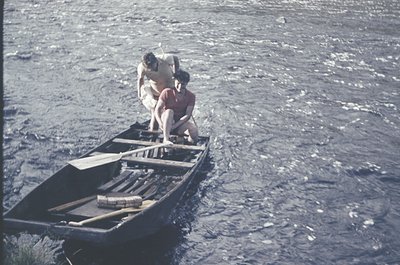 Vintage black-and-white boat scene: two individuals in a narrow wooden canoe, one kneeling to balance while the other stands....