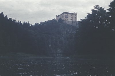 Historic castle perched atop rocky cliffside, surrounded by dense forest. Reflected in calm waters below, suggesting a lakesi...