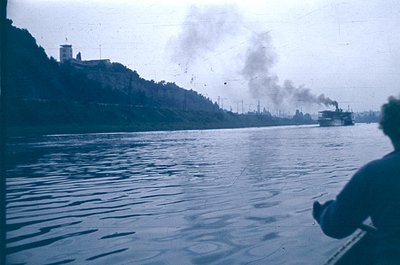 Vintage black-and-white photo of a steamboat emitting smoke on a river, with a hillside fortress and industrial structures in...