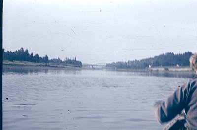 Vintage photo of a serene lake scene with partial view of a person’s arm holding a camera. Dense tree line frames the horizon...