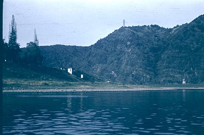 Vintage lake view with forested hills and sparse buildings. The serene reflection in calm waters suggests a remote, natural s...